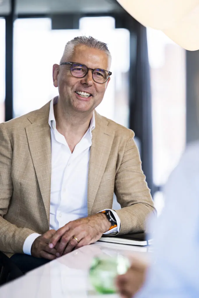 A smiling man with short, gray hair and glasses is seated at a table. He is wearing a light brown blazer over a white shirt. His hands are resting on the table, and he has a smartwatch on his wrist. In the foreground, a blurred hand is holding a green glass object, and a notebook is visible on the table. The background features large windows letting in natural light.