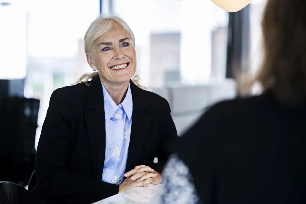 A woman with long, light-colored hair and a warm smile is seated at a table, wearing a black blazer over a light blue shirt. She appears engaged in conversation, with her hands clasped in front of her. The background shows a bright, modern office space with large windows.