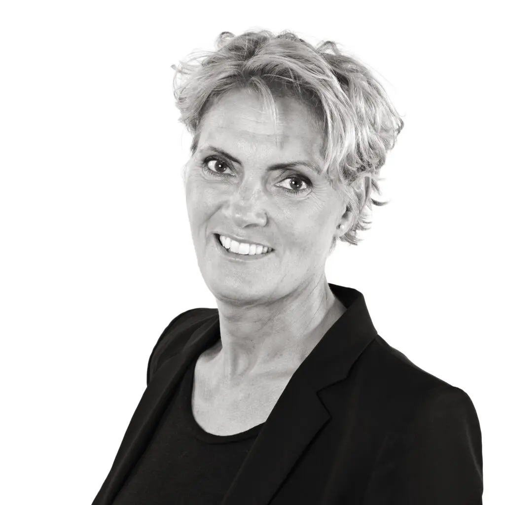 A woman with short, curly blonde hair smiles at the camera. She is wearing a black blazer over a black top. The background is white.