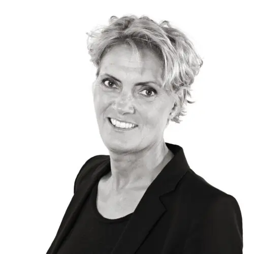 A woman with short, curly blonde hair smiles at the camera. She is wearing a black blazer over a black top. The background is white.