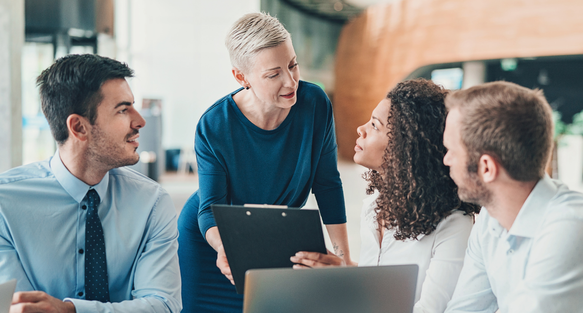 A group of four professionals is engaged in a discussion around a table. A woman with short, light hair is standing and holding a clipboard, addressing a woman with curly hair who is sitting and looking attentively. Two men, one with short dark hair and the other with light hair, are also seated at the table, listening to the conversation. The setting appears to be a modern office with natural light and wooden accents in the background.