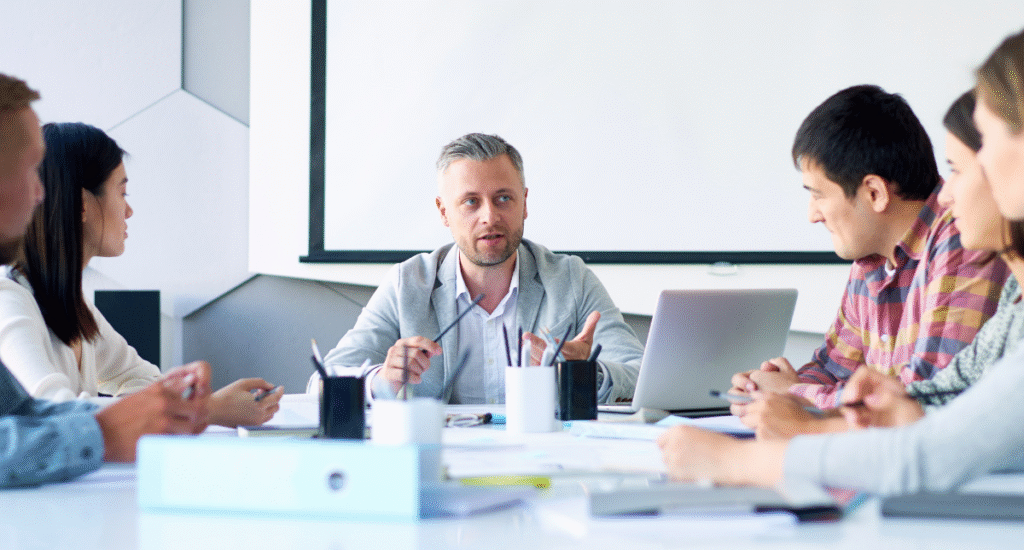 A group of six people is seated around a conference table. One man, wearing a light gray blazer, is speaking while holding a pen. He appears to be leading the discussion. A laptop is open in front of him, and there are various papers and notebooks on the table. The background features a large screen on the wall, which is currently blank. The group looks engaged in the conversation.