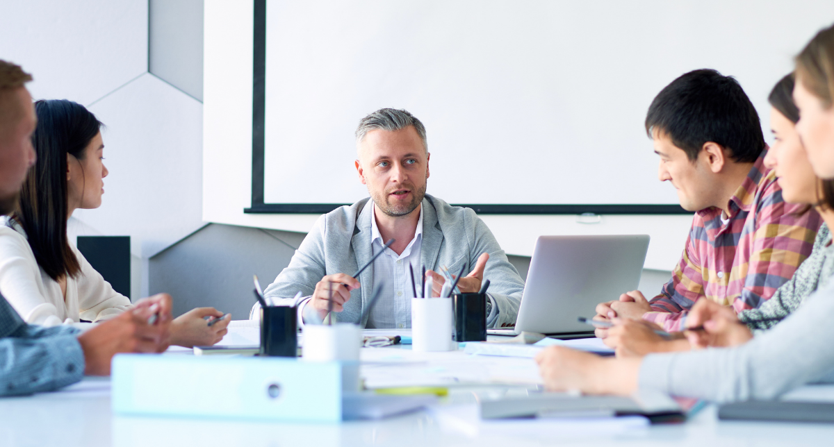 A group of six people is seated around a conference table. One man, wearing a light gray blazer, is speaking while holding a pen. He appears to be leading the discussion. A laptop is open in front of him, and there are various papers and notebooks on the table. The background features a large screen on the wall, which is currently blank. The group looks engaged in the conversation.