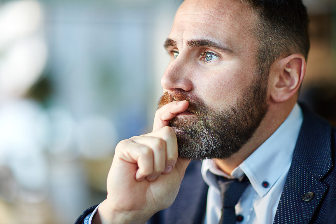 A man with a well-groomed beard and short hair sits thoughtfully, resting his finger on his lips. He is wearing a suit with a collared shirt and tie. The background is blurred, suggesting a professional or casual setting.