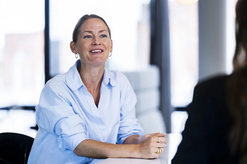 A woman with long, light brown hair, wearing a light blue button-up shirt, smiles while sitting at a table. Her hands are clasped together in front of her. The background features large windows with a blurred view of an outdoor setting, creating a bright and modern atmosphere. Another person is partially visible on the right, facing her.