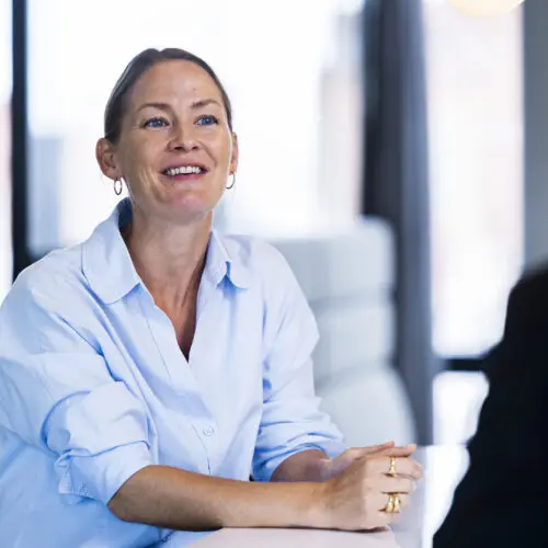 A woman with long, light brown hair, wearing a light blue button-up shirt, smiles while sitting at a table. Her hands are clasped together in front of her. The background features large windows with a blurred view of an outdoor setting, creating a bright and modern atmosphere. Another person is partially visible on the right, facing her.