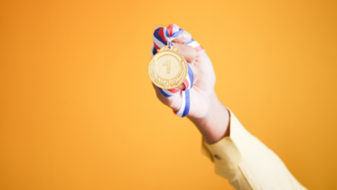 A hand holding a gold medal with a number "1" on it, attached to a red, white, and blue ribbon. The background is a solid orange color.