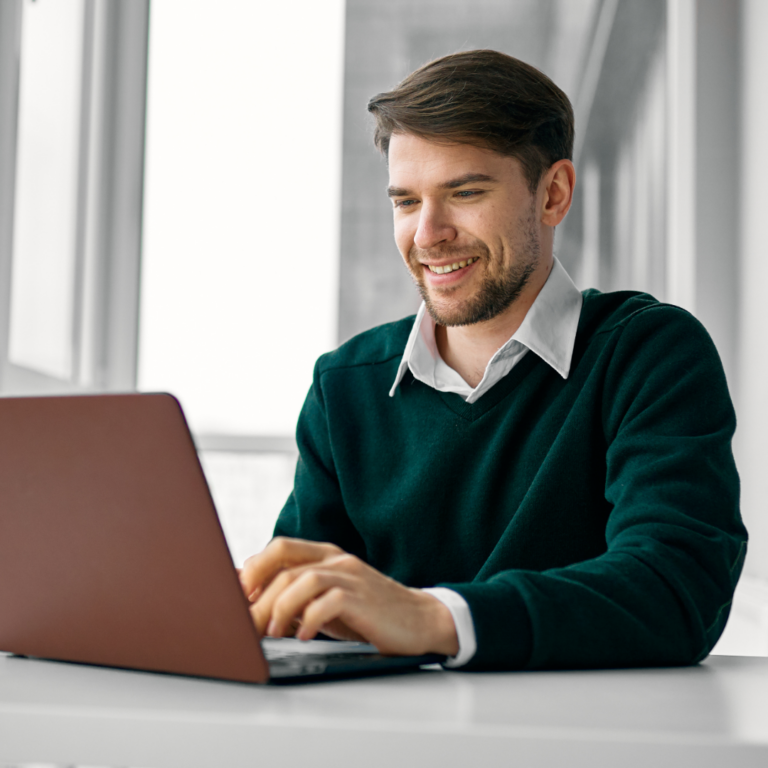 A young man with short brown hair and a beard is sitting at a white table, smiling as he types on a laptop. He is wearing a dark green sweater over a white collared shirt. The background features large windows with soft natural light.