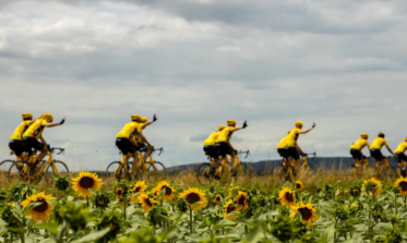 En gruppe cyklister i gule trøjer cykler langs en sti og vinker, mens de passerer. I forgrunden ses en mark med solsikker, hvor nogle blomster står i fuldt flor. Himlen er delvist skyet, hvilket skaber en lys udendørsscene.