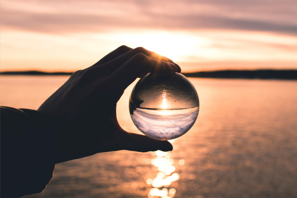 A hand holding a clear glass sphere in front of a sunset over water. The sphere reflects the sun and clouds, creating a distorted view of the scene. The background features a calm body of water with a soft golden light reflecting on its surface.