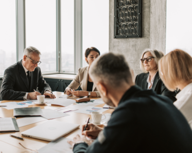 Ledergruppeudvikling. A group of five professionals is seated around a conference table in a modern office setting. They are engaged in discussion, with some taking notes and others looking at documents. The table is cluttered with papers displaying graphs and charts, a laptop, and coffee cups. Large windows in the background offer a view of the city skyline. The individuals are dressed in business attire, conveying a serious and collaborative atmosphere.