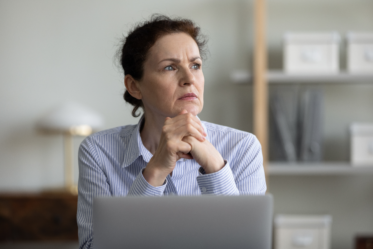 A woman with curly hair sits at a desk, looking thoughtfully into the distance. She has her hands clasped in front of her chin and is wearing a blue and white striped shirt. In the background, there are shelves with various storage boxes and a lamp. A laptop is open on the desk in front of her.