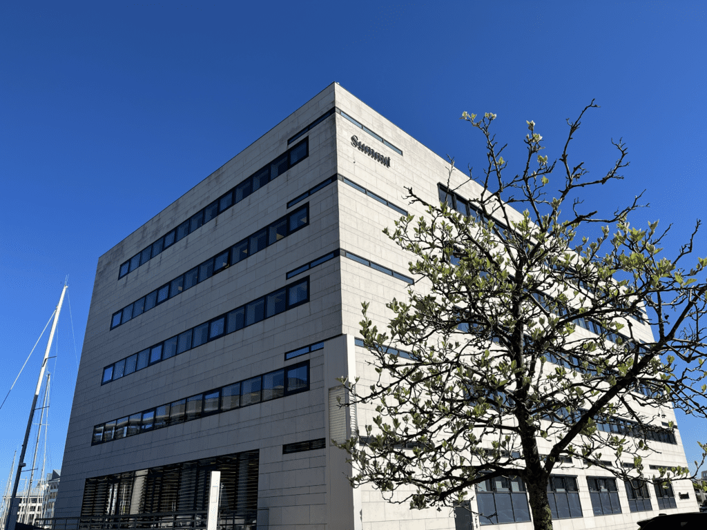 A modern, multi-story building with a light-colored façade and several windows, set against a clear blue sky. In the foreground, there is a tree with budding leaves. A sailboat is partially visible on the left side of the image.