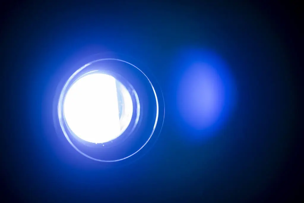 A close-up image of a bright circular light source against a dark background, with blue lighting effects surrounding it. The light appears to be emanating from a lens or projector.