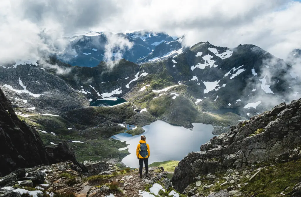 A person wearing a yellow jacket and a backpack stands on a rocky outcrop, overlooking a lush green valley filled with lakes and snow-capped mountains in the background. The sky is partly cloudy, with patches of blue visible among the clouds.