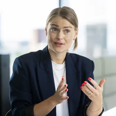 A person with light brown hair styled in a low ponytail is seated at a table, wearing a dark blazer over a white shirt. They are gesturing with their hands while speaking, displaying a focused expression. The background shows a bright, modern office space with large windows and a blurred cityscape outside.