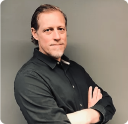 A man with light brown hair stands with his arms crossed, wearing a black button-up shirt. He is facing the camera against a neutral gray background. This image is associated with the "Past Events" page.
