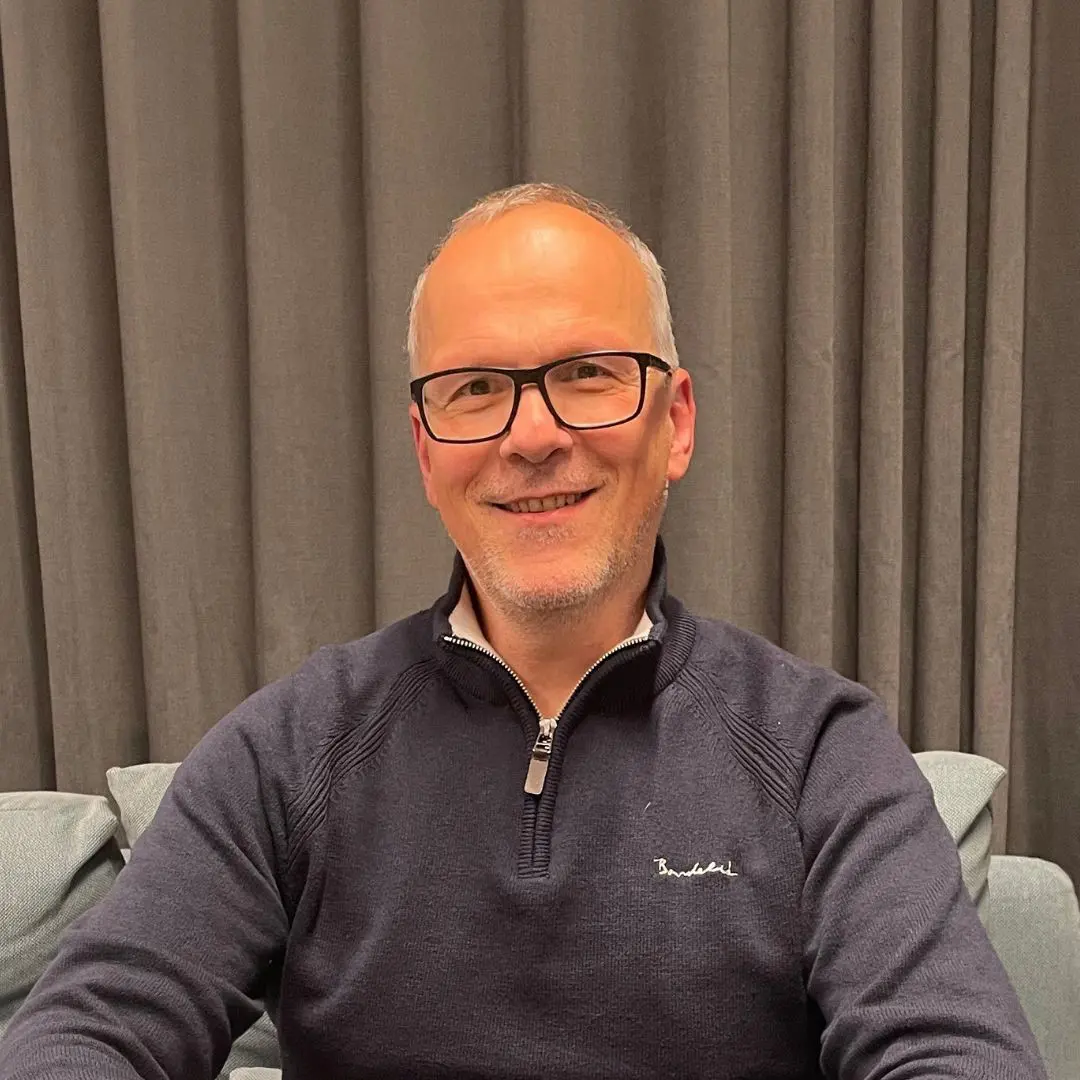 A smiling man with glasses is seated in front of a gray backdrop and a light blue couch. He is wearing a dark sweater with a zippered collar. The image appears on a page titled "Past Events."