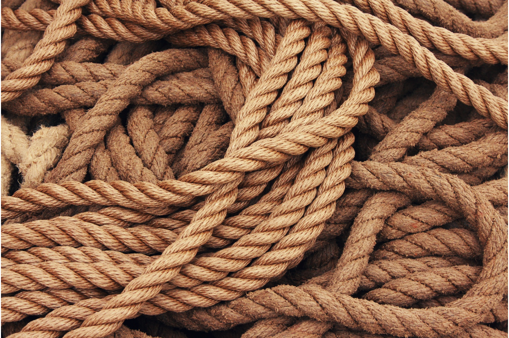 A close-up image of intertwined, thick, brown ropes arranged in a tangled pattern. The texture of the ropes is visible, showing their natural fibers and twists. The image evokes a sense of complexity, which can relate to the theme of "Dependability and People-Pleasing at Work."