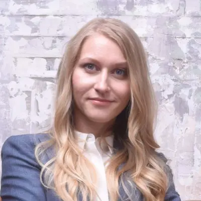 A woman with long, wavy blonde hair is standing with her arms crossed, wearing a dark blue blazer and a white blouse. She is looking directly at the camera with a neutral expression. The background features a textured, light-colored wall with a worn, artistic appearance.
