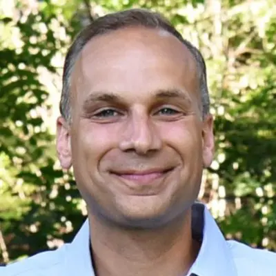 A smiling man with short, light brown hair and a light blue shirt is positioned in front of a blurred green background of trees. The image portrays a friendly and approachable demeanor.