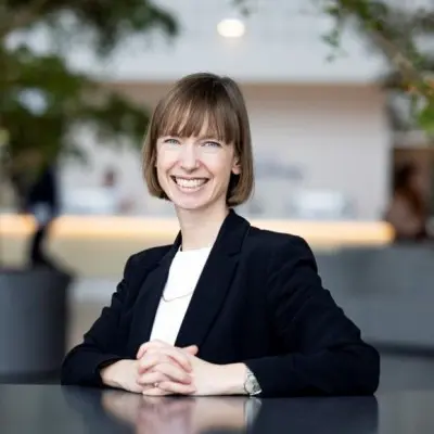 A woman with short brown hair smiles while sitting at a table with her hands clasped. She is wearing a black blazer over a white top. In the background, there are blurred outlines of indoor plants and a light-colored reception area. This image is featured on a page titled "Past Events."