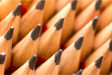 A close-up image of sharpened wooden pencils arranged closely together, with the pointed tips visible. The pencils have a light wooden body and dark graphite tips, creating a uniform and organized appearance. The background is slightly blurred, emphasizing the pencils in the foreground.