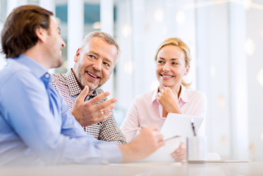 A group of three professionals is engaged in a conversation at a modern office setting. Two men and one woman are smiling and looking at each other, with one man gesturing while speaking. The woman is holding a tablet, and the atmosphere appears collaborative and friendly, with soft lighting and a bright, airy background.