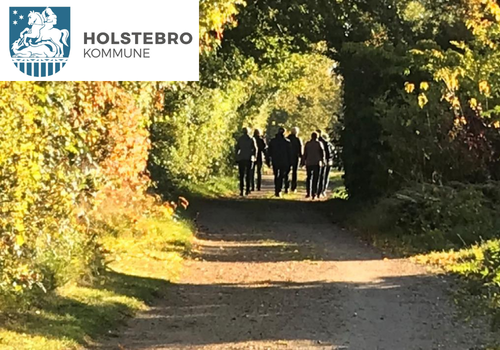 En gruppe på fem personer går ad en træomkranset sti, omgivet af grønne omgivelser og efterårsfarver. Scenen er godt oplyst, hvilket tyder på en solrig dag. Holstebro Kommunes logo vises i øverste venstre hjørne.