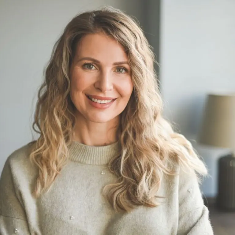 A woman with long, wavy blonde hair smiles warmly at the camera. She is wearing a light gray sweater adorned with small embellishments. The background features a softly lit interior with a lamp and a piece of furniture partially visible.