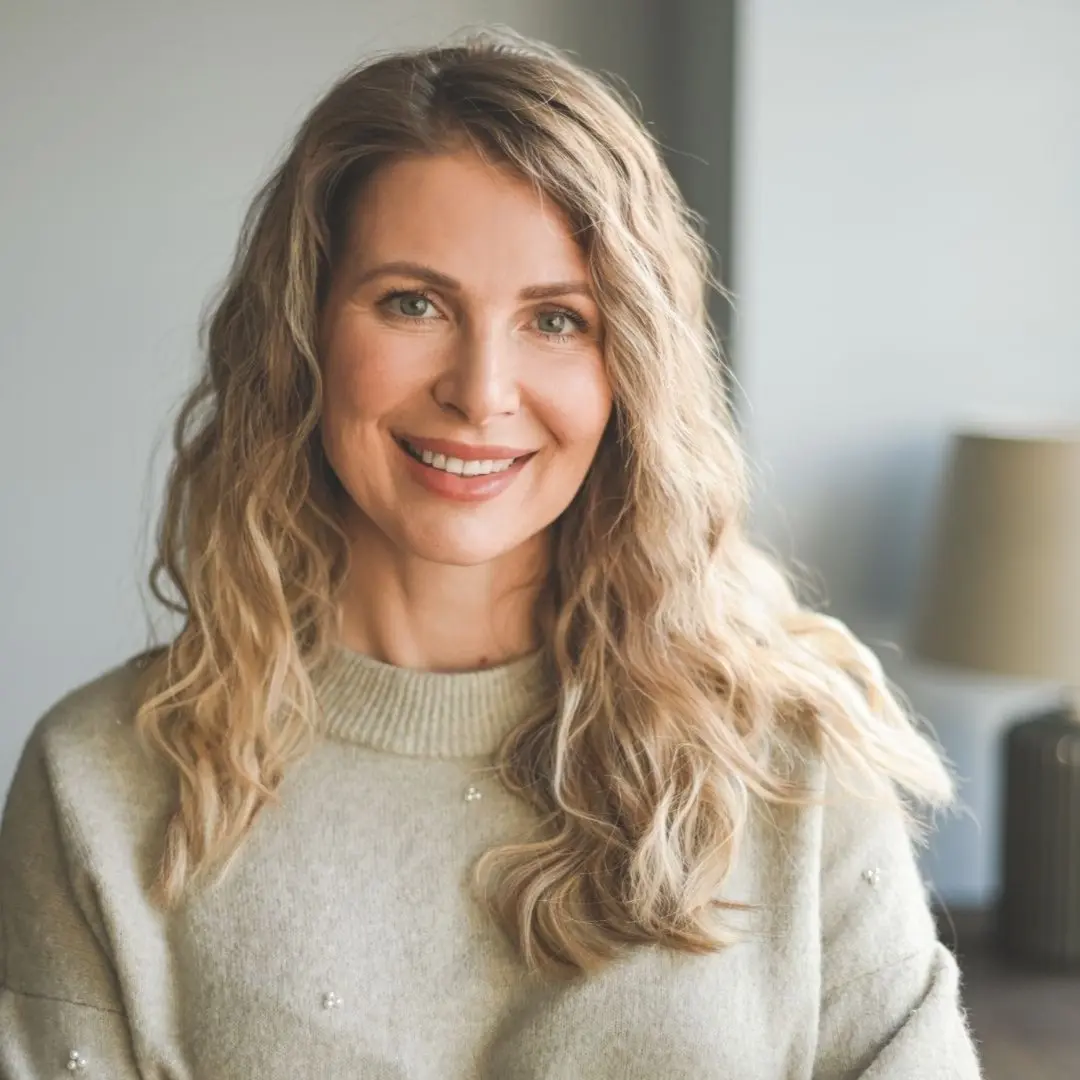 A woman with long, wavy blonde hair smiles warmly at the camera. She is wearing a light gray sweater adorned with small embellishments. The background features a softly lit interior with a lamp and a piece of furniture partially visible.