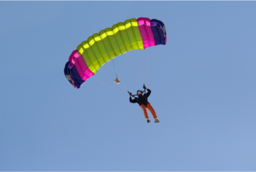 A person is parachuting from the sky, suspended beneath a brightly colored parachute with green, pink, and purple sections. The individual is wearing an orange jumpsuit and a black jacket, and is descending against a clear blue sky. This image relates to the theme of risk-taking in leadership.