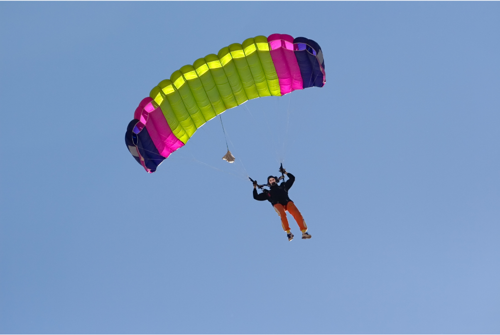 A person is parachuting from the sky, suspended beneath a brightly colored parachute with green, pink, and purple sections. The individual is wearing an orange jumpsuit and a black jacket, and is descending against a clear blue sky. This image relates to the theme of risk-taking in leadership.