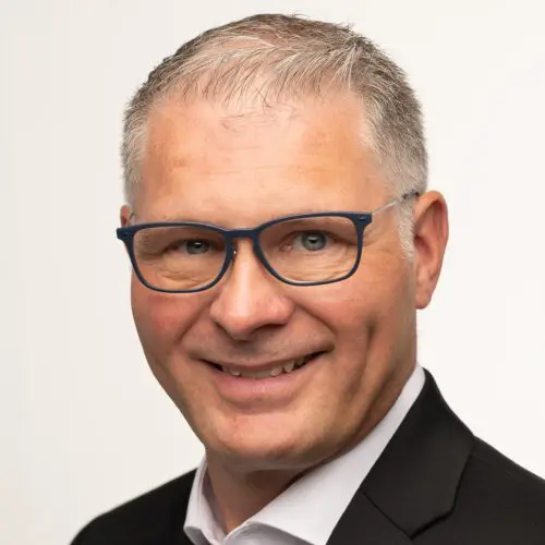 A close-up portrait of Hans Fogh Laustsen, featuring a man with short, light gray hair and wearing blue glasses. He has a friendly smile and is dressed in a black suit with a white shirt. The background is plain white.