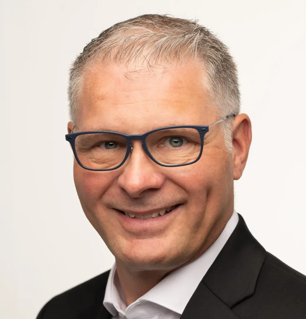 A close-up portrait of Hans Fogh Laustsen, featuring a man with short, light gray hair and wearing blue glasses. He has a friendly smile and is dressed in a black suit with a white shirt. The background is plain white.