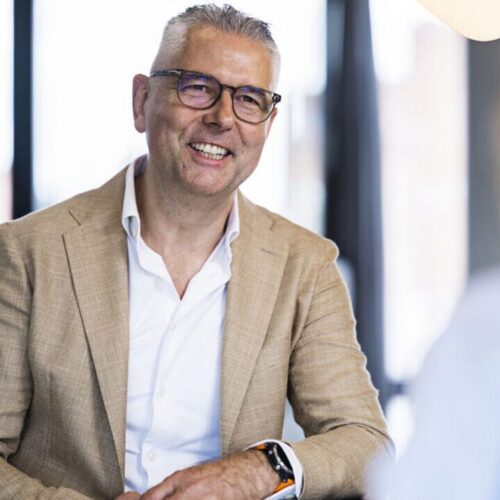 A smiling man with short, gray hair and glasses is seated at a table. He is wearing a light brown blazer over a white shirt. His hands are resting on the table, and he has a smartwatch on his wrist. In the foreground, a blurred hand is holding a green glass object, and a notebook is visible on the table. The background features large windows letting in natural light.