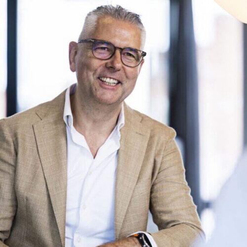 A smiling man with short, gray hair and glasses is seated at a table. He is wearing a light brown blazer over a white shirt. His hands are resting on the table, and he has a smartwatch on his wrist. In the foreground, a blurred hand is holding a green glass object, and a notebook is visible on the table. The background features large windows letting in natural light.