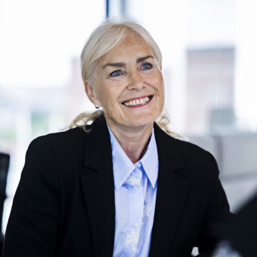 A woman with long, light-colored hair and a warm smile is seated at a table, wearing a black blazer over a light blue shirt. She appears engaged in conversation, with her hands clasped in front of her. The background shows a bright, modern office space with large windows.