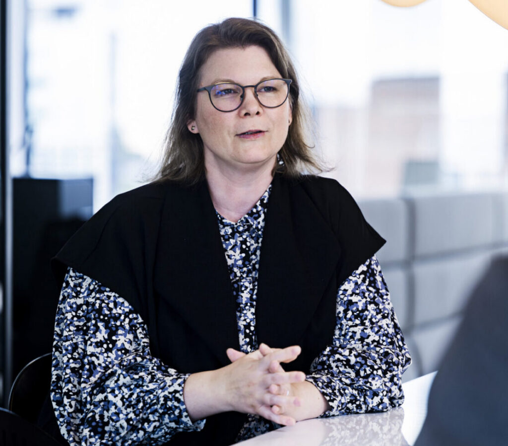 A woman with shoulder-length brown hair and glasses is seated at a table, engaging in conversation. She wears a black vest over a patterned blouse featuring shades of blue and white. The background includes large windows with a cityscape visible outside. The focus is on her upper body and hands, which are clasped together. Another person, partially visible, appears to be listening to her.