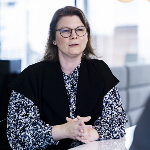 A woman with shoulder-length brown hair and glasses is seated at a table, engaging in conversation. She wears a black vest over a patterned blouse featuring shades of blue and white. The background includes large windows with a cityscape visible outside. The focus is on her upper body and hands, which are clasped together. Another person, partially visible, appears to be listening to her.
