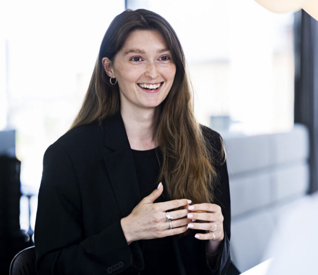 A woman with long, wavy brown hair, wearing a black blazer, is smiling and speaking while sitting at a table. She gestures with her hands, indicating engagement in conversation. In the background, another woman with her back to the camera is partially visible, and the setting appears to be a bright, modern office space.