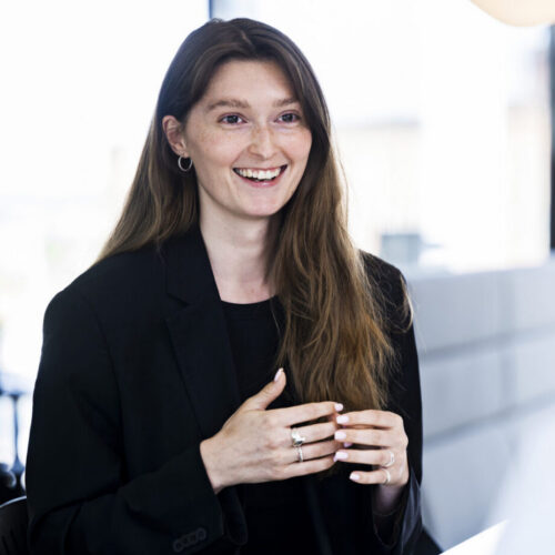 A woman with long, wavy brown hair, wearing a black blazer, is smiling and speaking while sitting at a table. She gestures with her hands, indicating engagement in conversation. In the background, another woman with her back to the camera is partially visible, and the setting appears to be a bright, modern office space.
