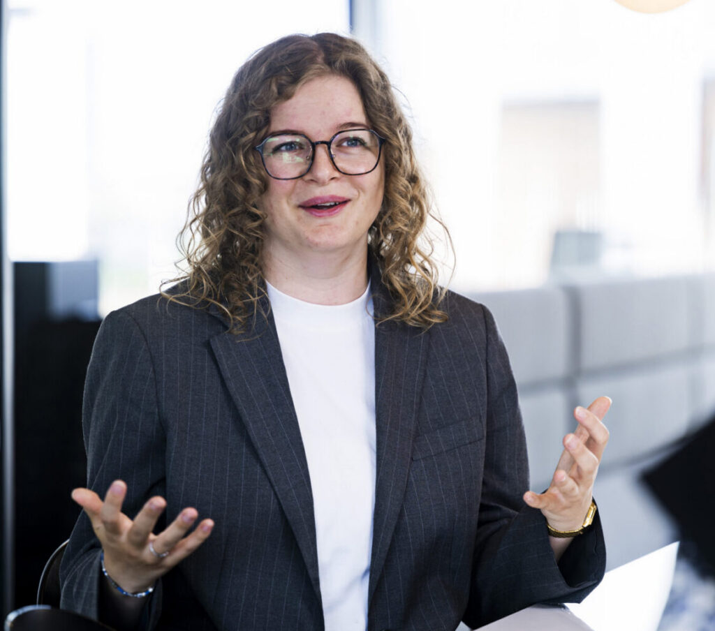 A woman with curly hair and glasses is sitting at a table, wearing a dark blazer over a white shirt. She appears to be engaged in conversation, using her hands for emphasis. The background features modern office decor with large windows and soft lighting.