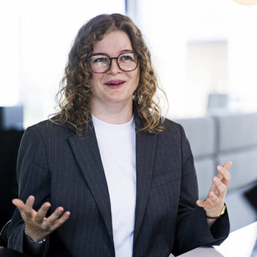 A woman with curly hair and glasses is sitting at a table, wearing a dark blazer over a white shirt. She appears to be engaged in conversation, using her hands for emphasis. The background features modern office decor with large windows and soft lighting.