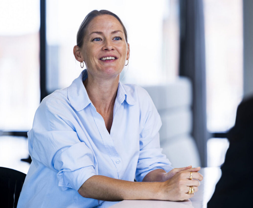 A woman with long, light brown hair, wearing a light blue button-up shirt, smiles while sitting at a table. Her hands are clasped together in front of her. The background features large windows with a blurred view of an outdoor setting, creating a bright and modern atmosphere. Another person is partially visible on the right, facing her.