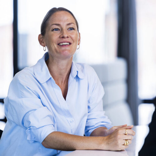 A woman with long, light brown hair, wearing a light blue button-up shirt, smiles while sitting at a table. Her hands are clasped together in front of her. The background features large windows with a blurred view of an outdoor setting, creating a bright and modern atmosphere. Another person is partially visible on the right, facing her.