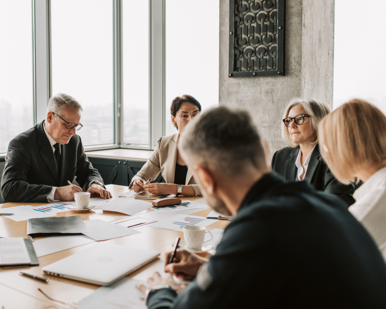 Ledergruppeudvikling. A group of five professionals is seated around a conference table in a modern office setting. They are engaged in discussion, with some taking notes and others looking at documents. The table is cluttered with papers displaying graphs and charts, a laptop, and coffee cups. Large windows in the background offer a view of the city skyline. The individuals are dressed in business attire, conveying a serious and collaborative atmosphere.