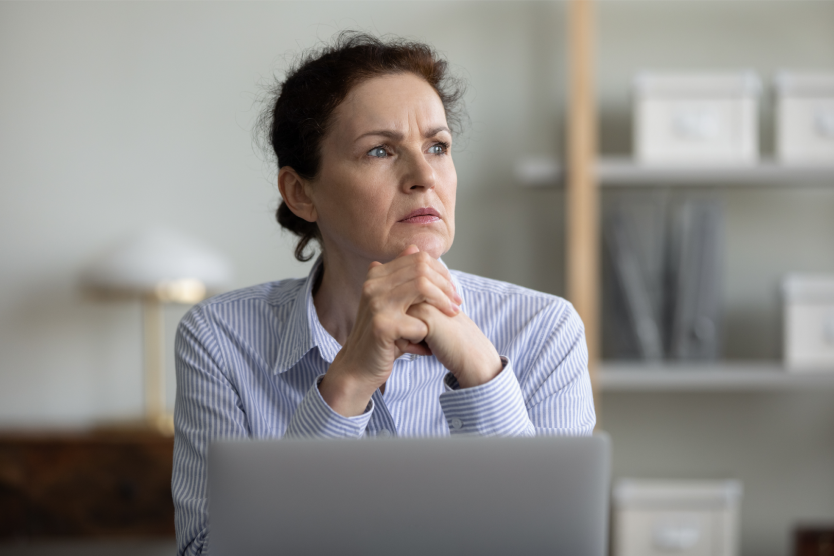 A woman with curly hair sits at a desk, looking thoughtfully into the distance. She has her hands clasped in front of her chin and is wearing a blue and white striped shirt. In the background, there are shelves with various storage boxes and a lamp. A laptop is open on the desk in front of her.