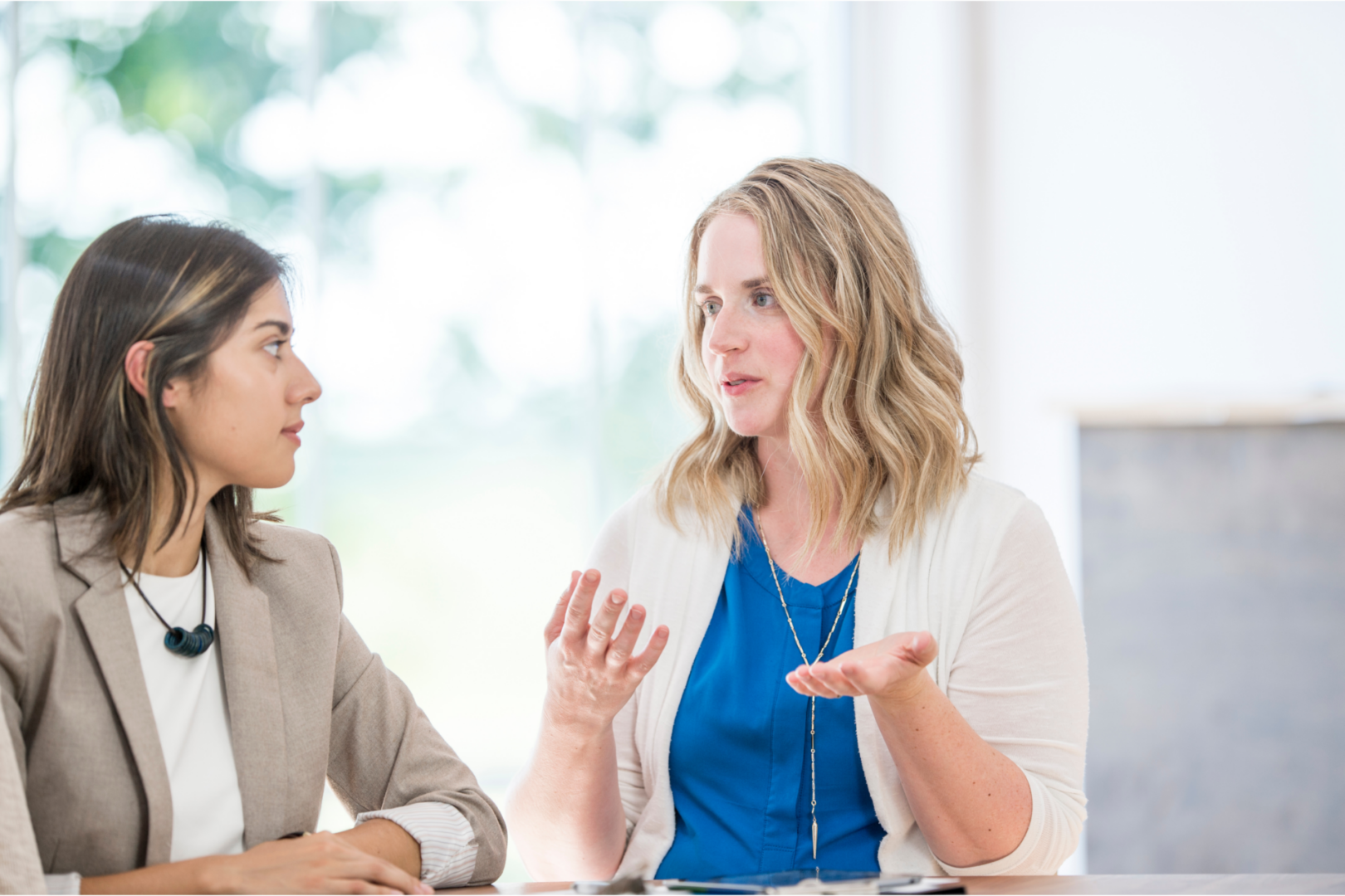 Two women are engaged in a conversation at a table. One woman, with wavy blonde hair, is speaking and using hand gestures to emphasize her points. The other woman, with dark hair, is listening attentively. The background features large windows with natural light coming in.